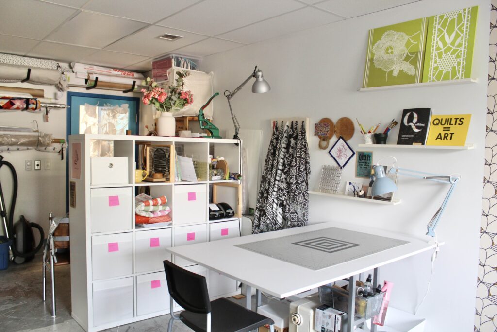 A view of a sewing studio with a large cutting table in the center, and a wall of shelves 