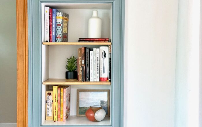 A kitchen nook in the wall displaying three shelves of cookbooks with other decorative items.