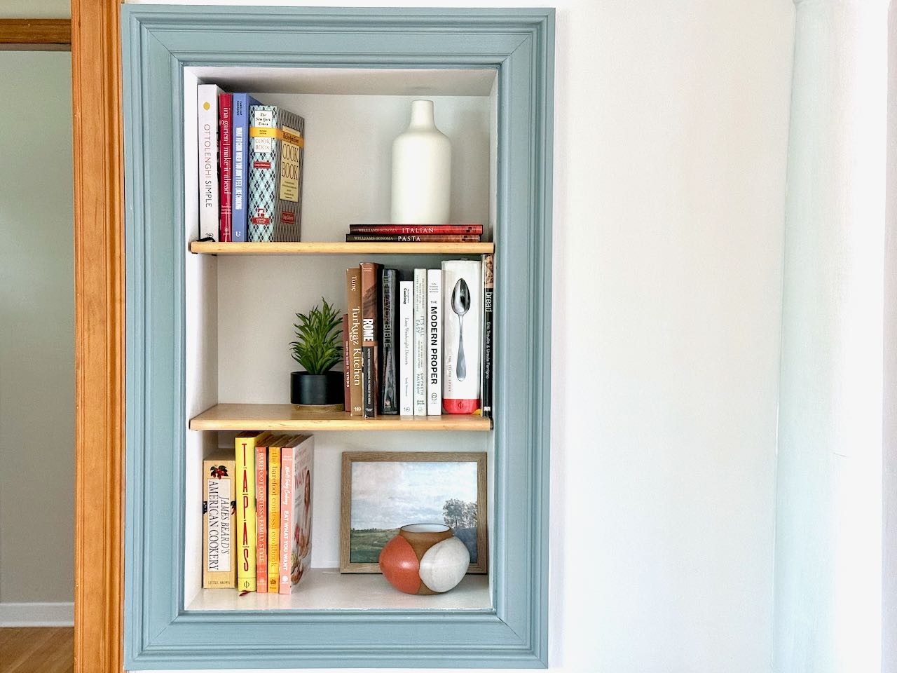 A kitchen nook in the wall displaying three shelves of cookbooks with other decorative items.