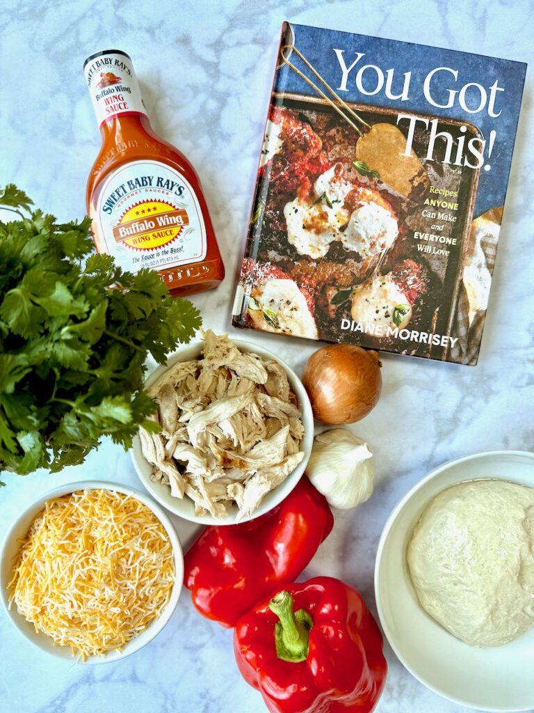 You Got This! Cookbook next to cilantro, sweet baby rays buffalo sauce rotisserie chicken shredded onion garlic red peppers pizza dough and Mexican shredded cheese