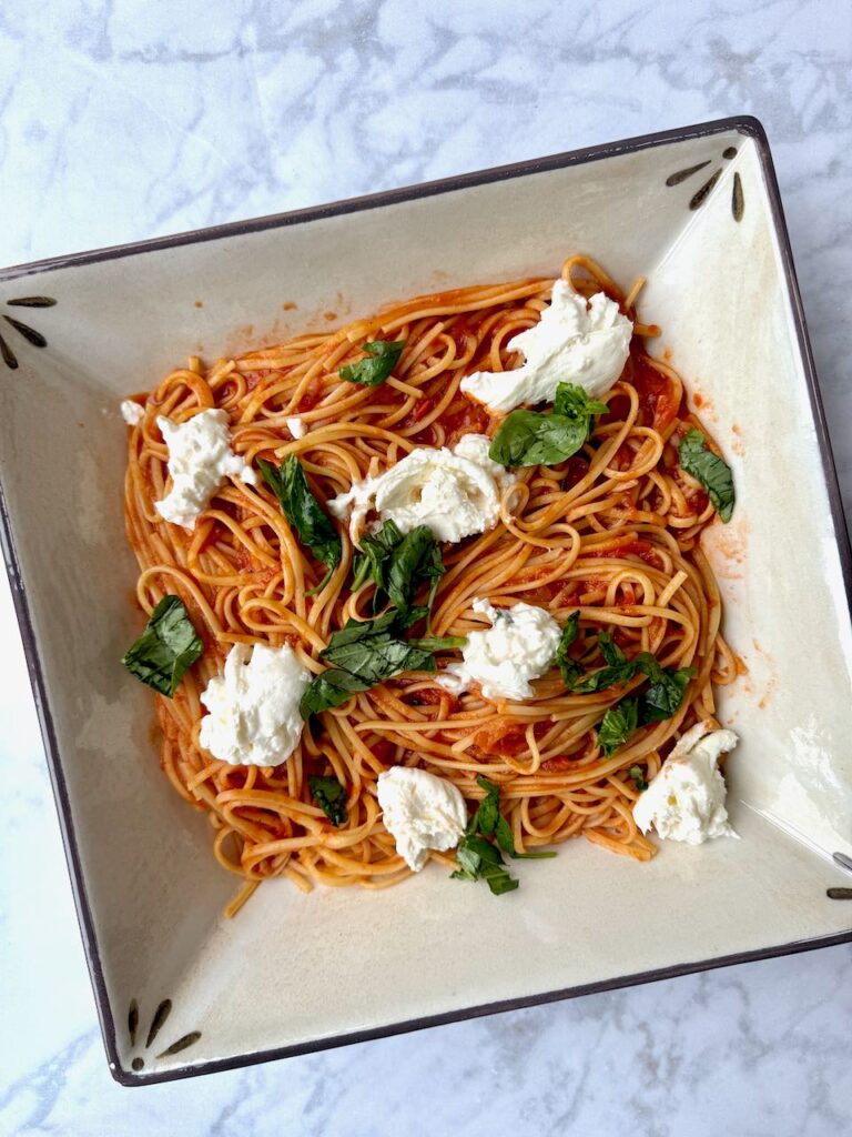 A square bowl of pasta with tomato sauce chunks of burrata and basil
