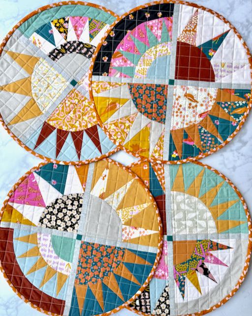 Four circular quilted placemats arranged in a grid, each featuring different color combinations of the same sunburst pattern with radiating fabric rays and orange polka dot binding.