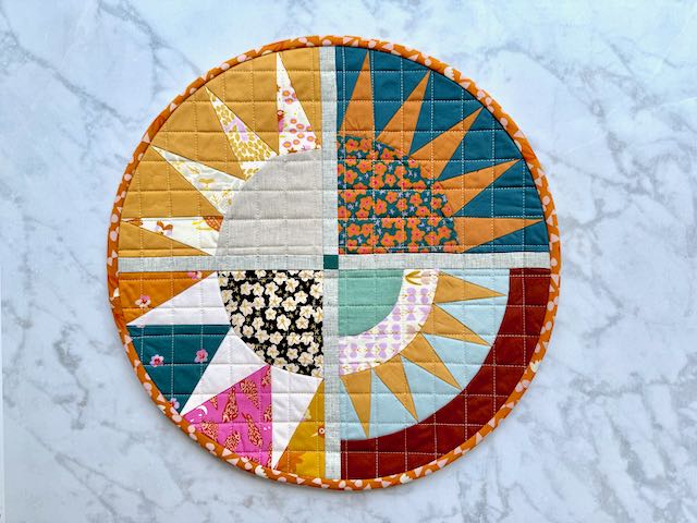 Another version of the same circular quilted sunburst placemat, showing different fabric combinations in bright colors including teal, pale blue, gold, orange, and teal, with the same orange patterned binding.