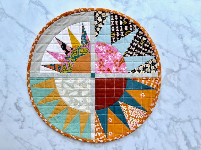 Another version of the same circular quilted sunburst placemat, showing different fabric combinations in autumn colors including seafoam green, orange, and teal, with the same orange patterned binding.
