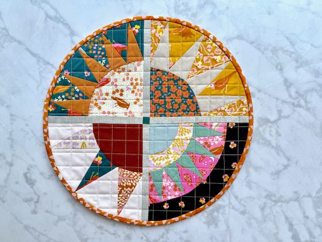 Another version of the same circular quilted sunburst placemat, showing different fabric combinations in autumn colors including rust, golden yellow, and teal, with the same orange patterned binding.