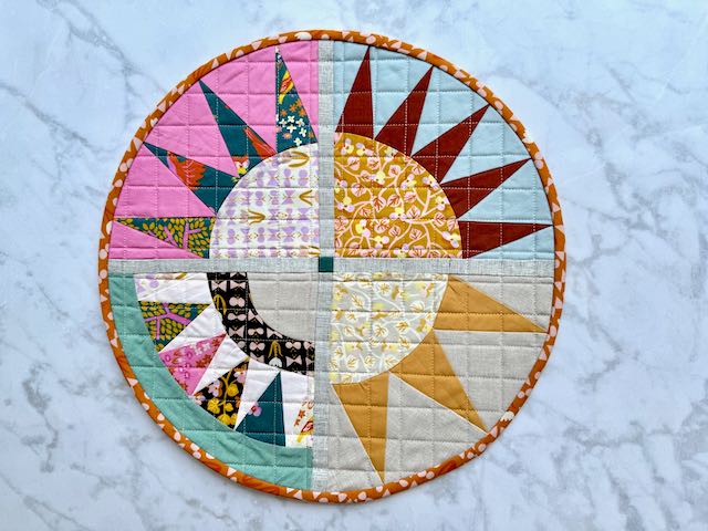 Circular quilted wall hanging with sunburst pattern in four quadrants, featuring colorful fabric rays radiating from the center against pink and blue backgrounds, finished with orange printed binding.