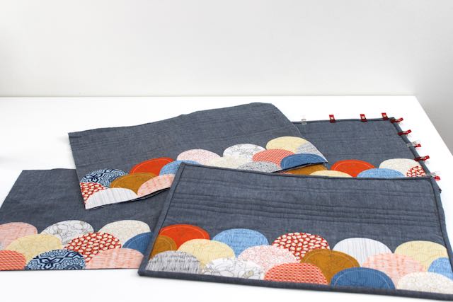 The fronts of rectangular placemats with clamshell scallops in an assortment of fabrics by Carolyn friedlander in oranges blues and white on a denim blue background.