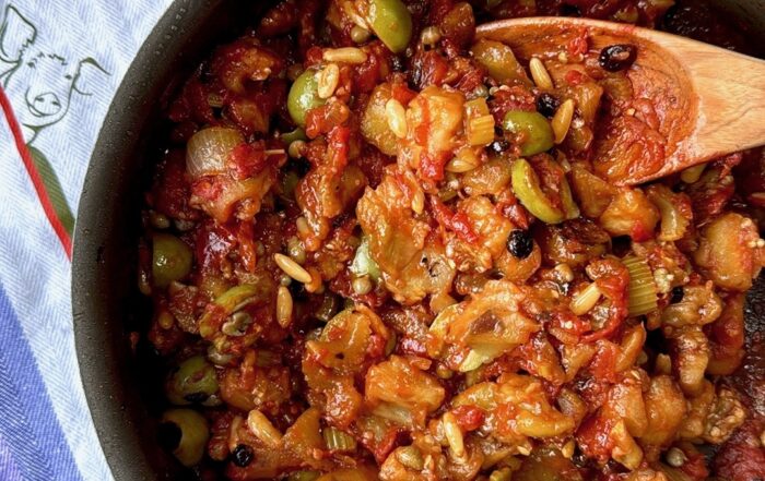 Close-up detail shot of caponata in the skillet showing individual ingredients like pine nuts, green olives, diced eggplant, and tomatoes glistening in olive oil with a wooden spoon.
