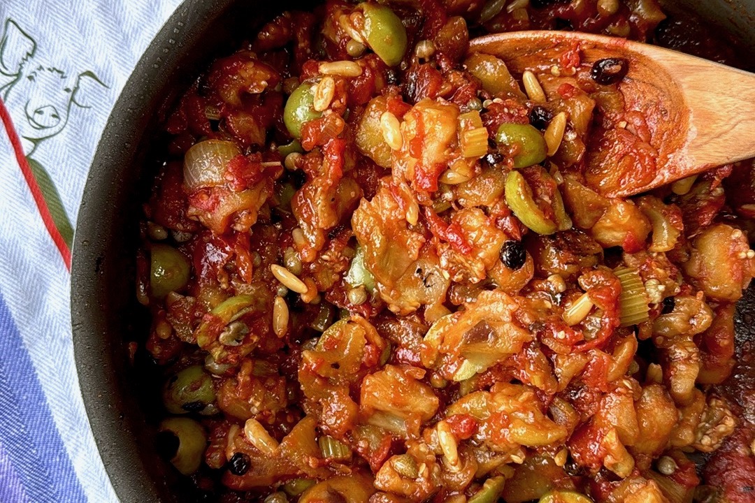 Close-up detail shot of caponata in the skillet showing individual ingredients like pine nuts, green olives, diced eggplant, and tomatoes glistening in olive oil with a wooden spoon.